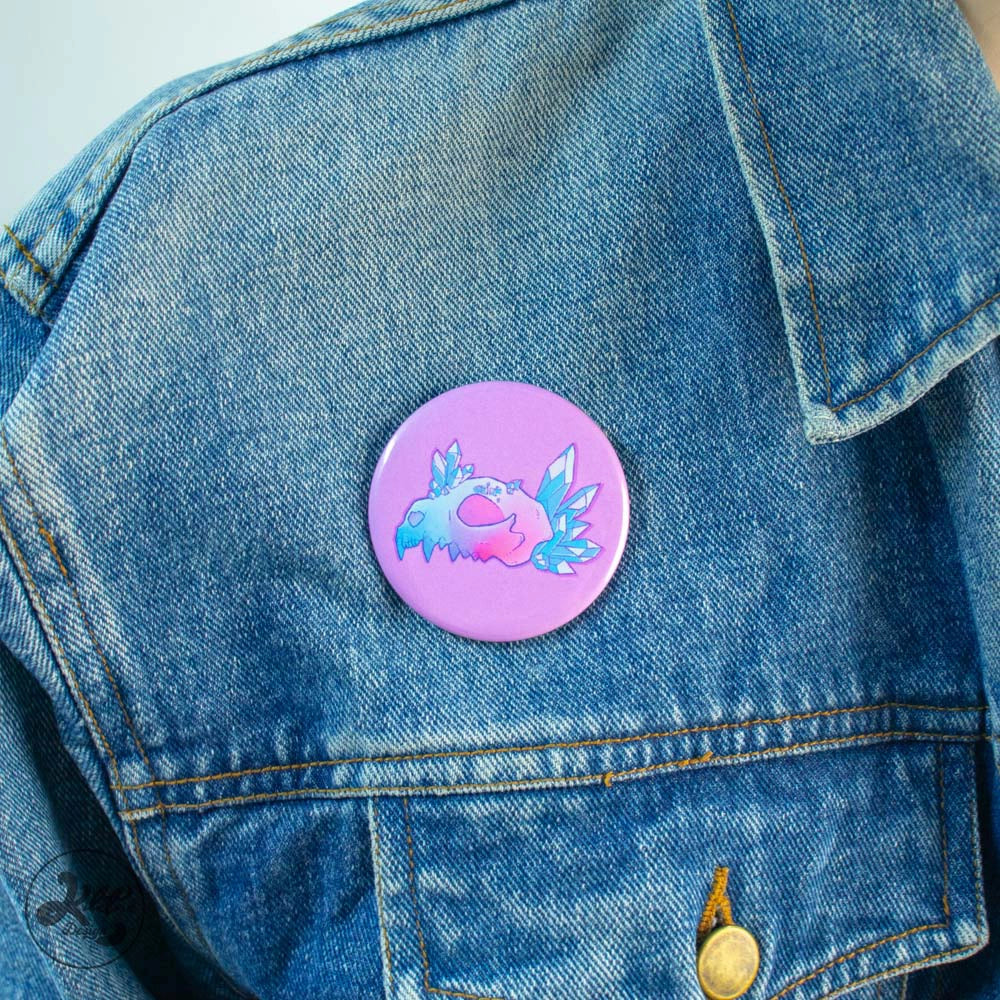 59mm button pin by Kilatora Design. Pink color and blue crystals next to pink and blue wolf skull. In the photo, the pin is attached to a denim jacket.