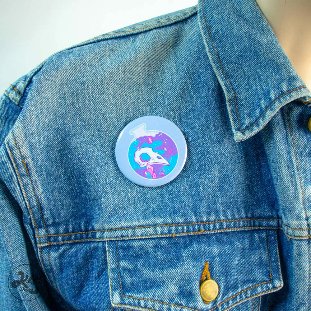 59mm button pin by Kilatora Design. Light blue color with a blue and purple round bottle with a bird's skull and pink crystals inside. In the photo, the pin is attached to a denim jacket.