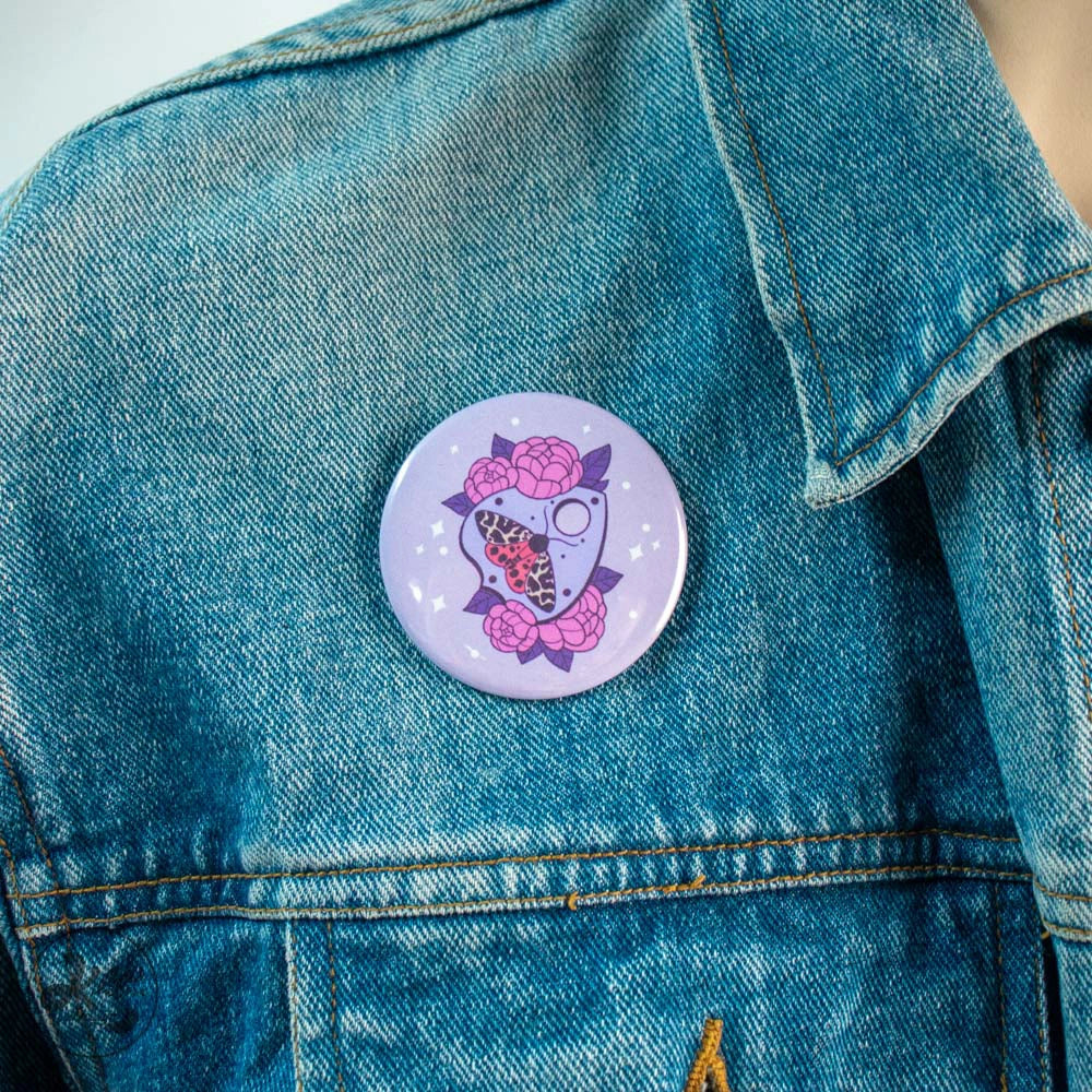 59mm button pin by Kilatora Design. Light purple color with purple ouija, a moth with white and orange wings and lilac flowers. In the photo, the pin is attached to a denim jacket.
