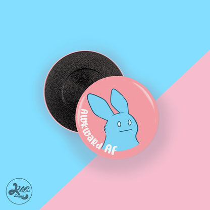 38mm button pin by Kilatora Design. Warm pink color with a blue bunny and text "awkward af".