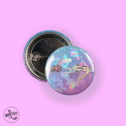 38mm button pin by Kilatora Design. Blue and purple color with broken glass holographic effect. A gray cat's paw reaches for a skeleton's hand.
