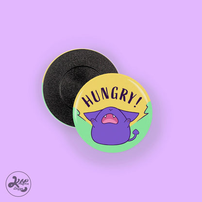 38mm button pin by Kilatora Design. Green pin with purple Tora and text  "hungry".