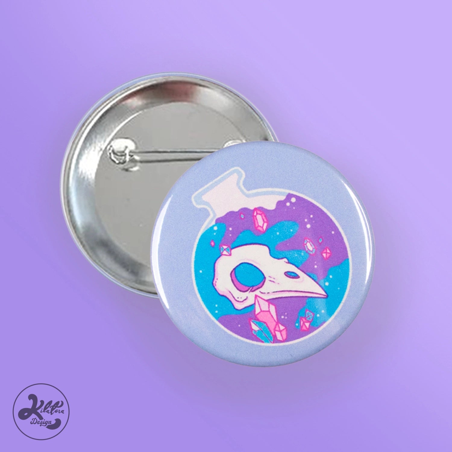 59mm button pin by Kilatora Design. Light blue color with a blue and purple round bottle with a bird's skull and pink crystals inside.