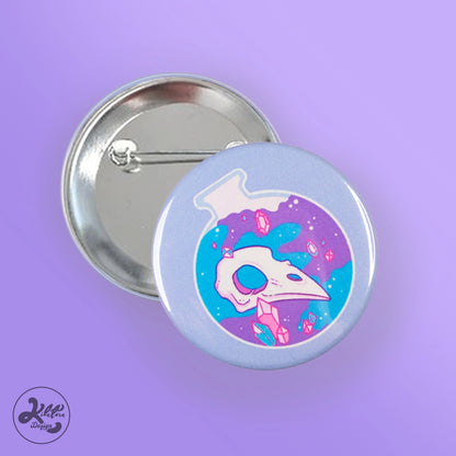 59mm button pin by Kilatora Design. Light blue color with a blue and purple round bottle with a bird's skull and pink crystals inside.