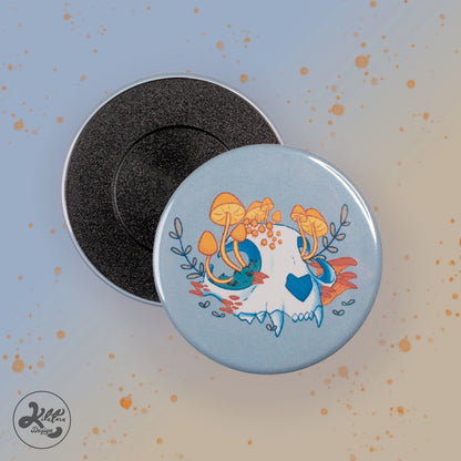 59 mm magnet by Kilatora Design. Light grey color with a white cat skull with yellow mushrooms and plants growing from it.